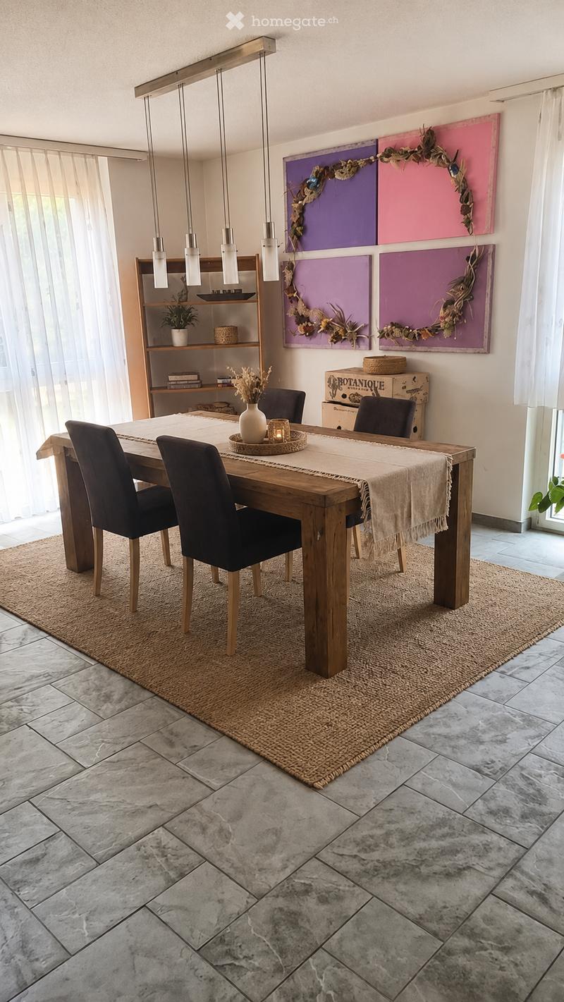 Dining room with a wooden dining table, four black chairs, a beige table runner, a white vase with dried flowers, a shelf with books and a small plant, and colorful wall art.