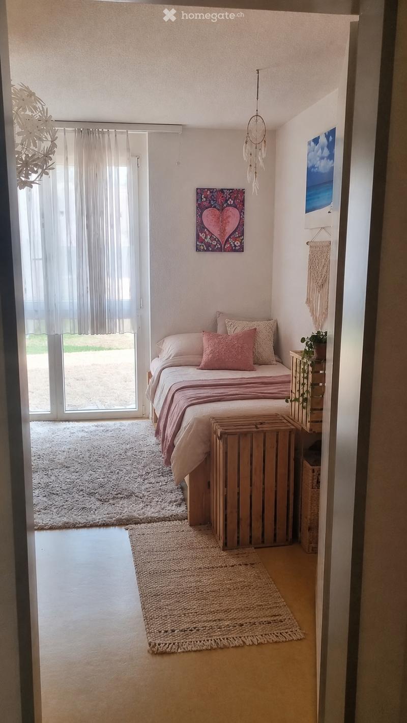 Cozy bedroom with a double bed, pink bedding, nightstand, potted plant, and artwork on the walls. The room features a large window with sheer curtains, a fluffy white rug, and a woven floor rug.