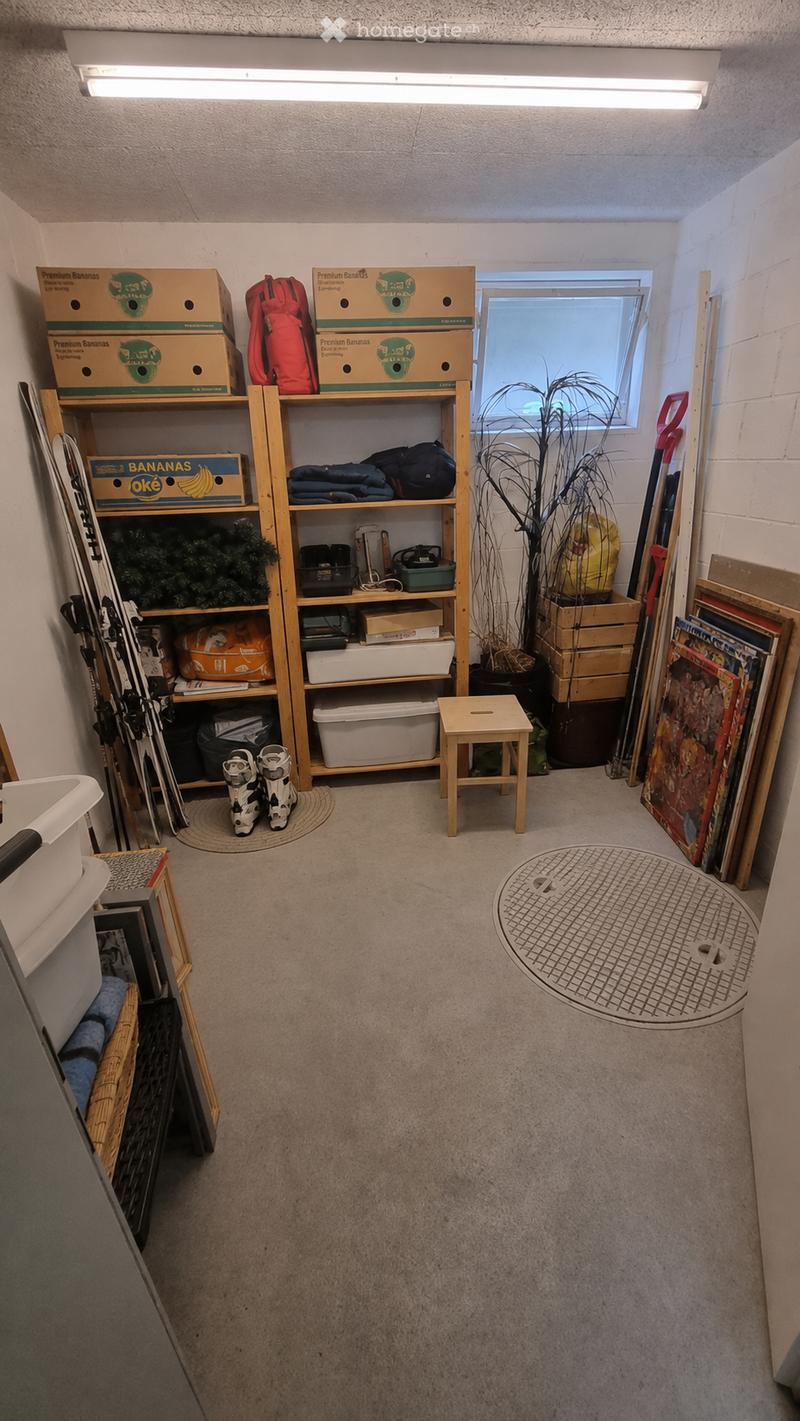 The image shows a storage room with a wooden shelf holding various items, including storage boxes, a backpack, skiis, a snowboard, and a potted plant. A small wooden stool is placed in the center of the room, and there are additional items such as paintin