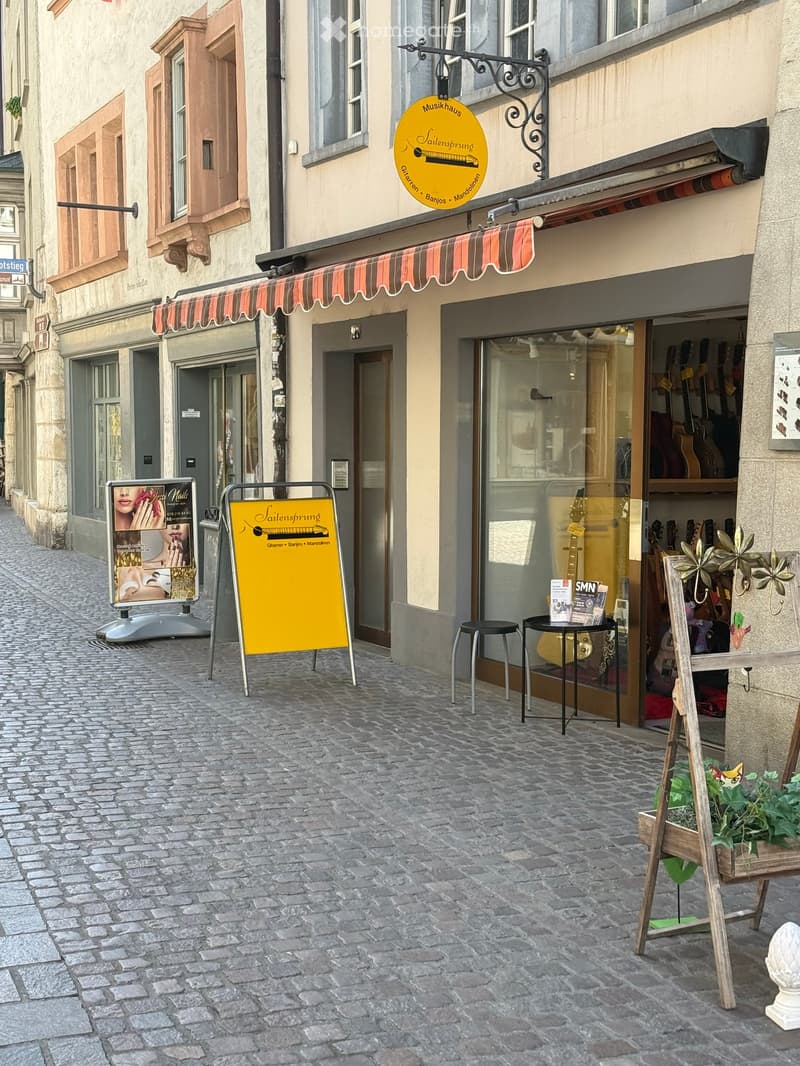 A narrow cobblestone street lined with historic buildings, featuring a storefront with a striped awning, a yellow signboard, and a small outdoor seating area with a table and chairs.