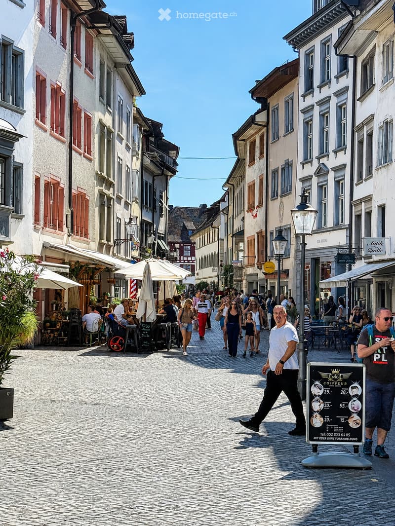 A bustling cobblestone street lined with historic buildings, outdoor seating areas, and pedestrians. The street is adorned with street lamps, signage, and a sign advertising a coffee bar.