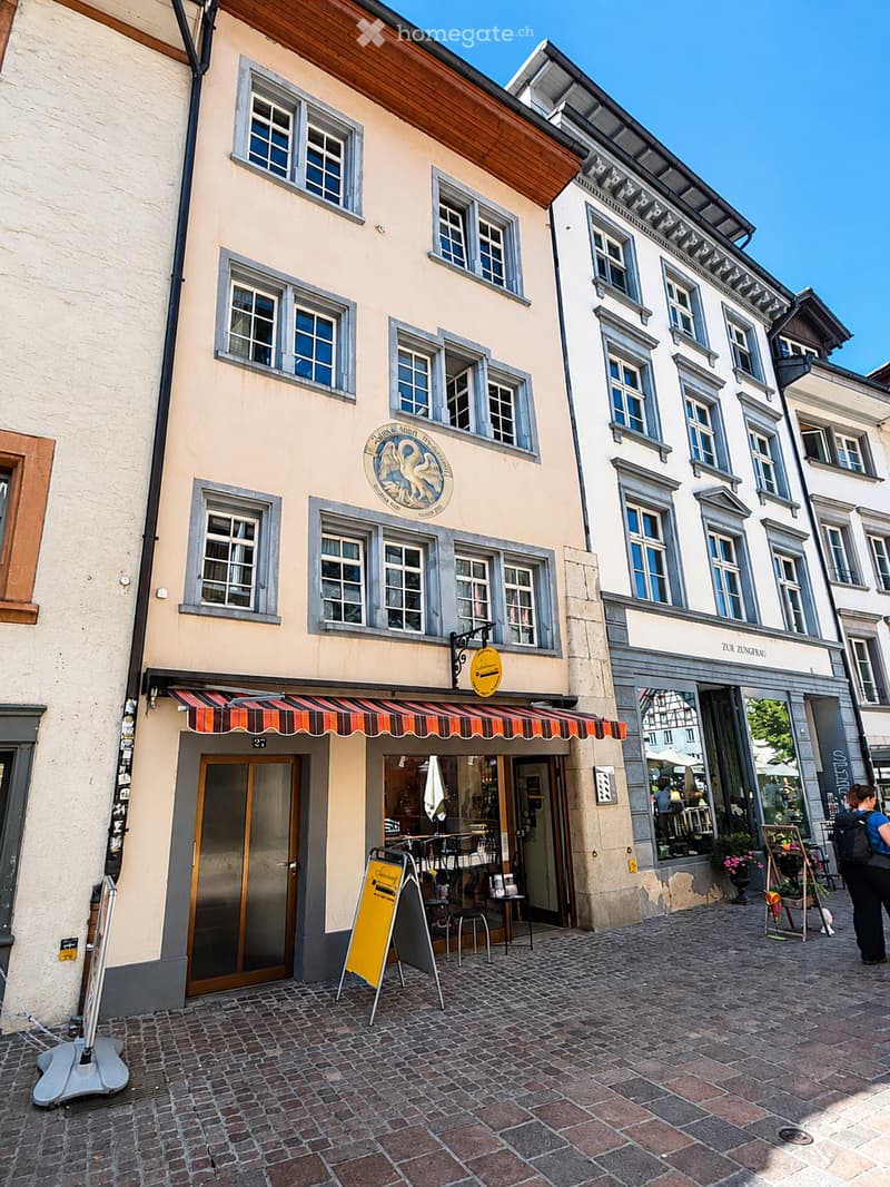 A multi-story building with a light-colored facade, multiple windows, and a striped awning over the entrance. The ground floor features a shop with visible tables and chairs inside, and a yellow signboard outside.