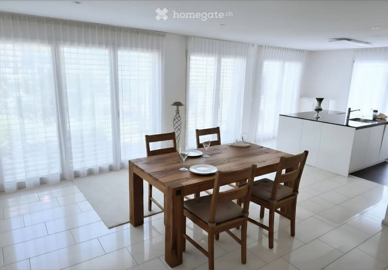 Bright dining area with a wooden dining table, four chairs, wine glasses, plates, and a modern kitchen area with white cabinets, a sink, and a vase on the counter.