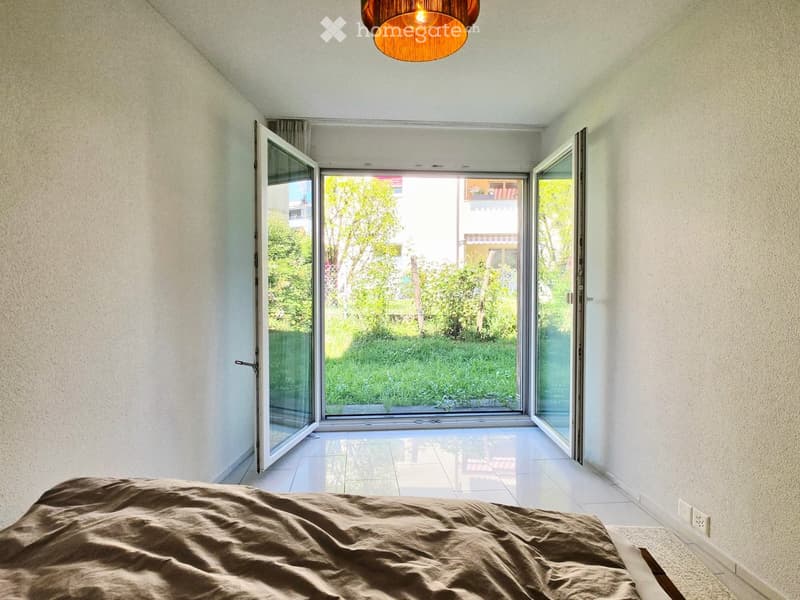 Modern bedroom with a large bed, white tiled floor, large glass doors opening to a garden view.