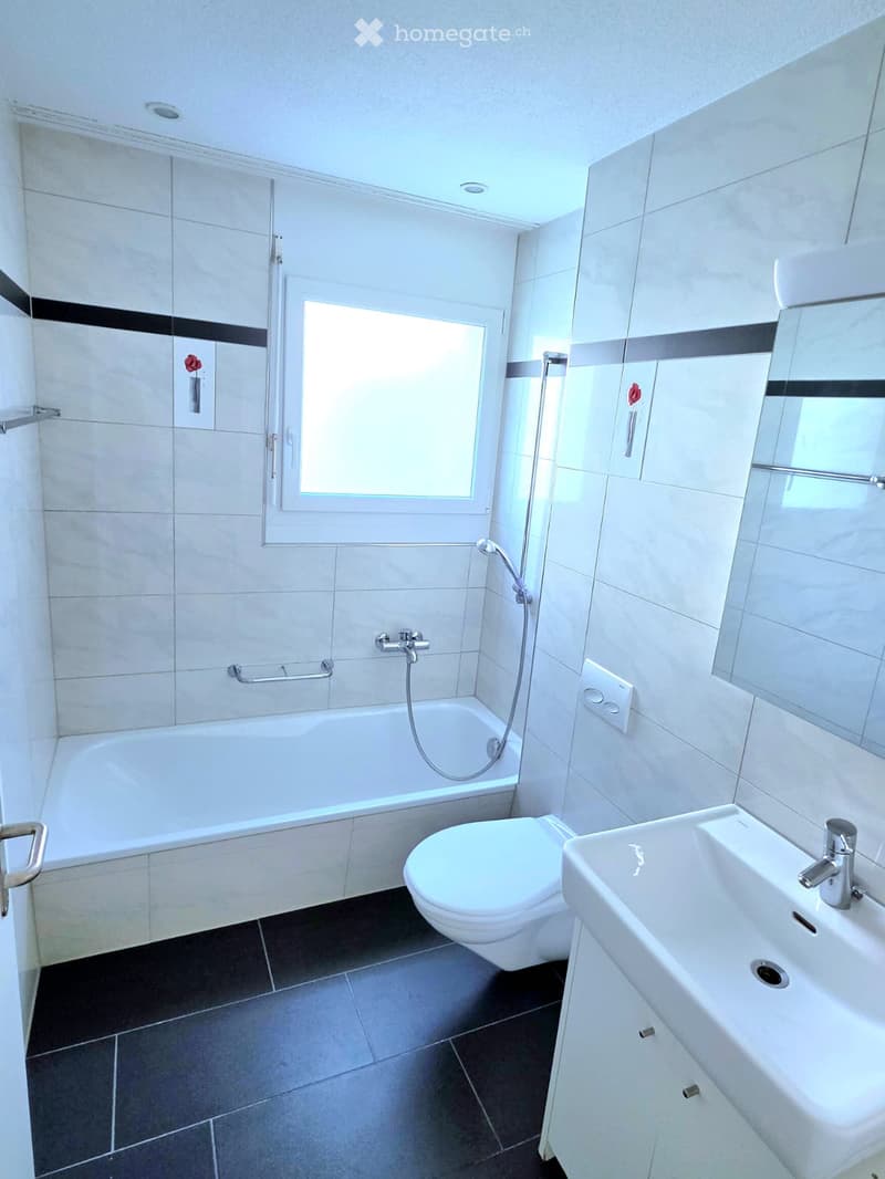 Modern bathroom with white tiles, bathtub, toilet, sink, and shower hose. Features a window and a small decorative flower on the wall.