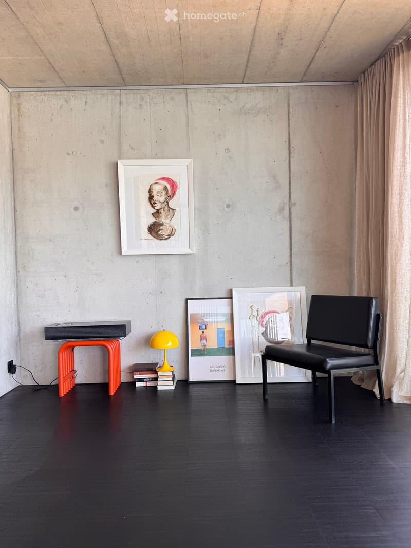 Modern, minimalist room with dark wooden flooring, concrete walls, and large picture frames. Features a black leather chair, orange stool, yellow lamp, and a small table with books.