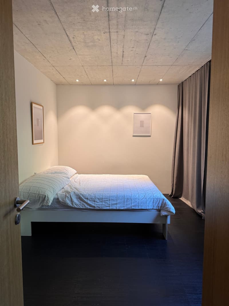 Minimalist bedroom with a single bed, pillows, and a dark wooden floor. Features a large window with curtains and two picture frames on the walls.
