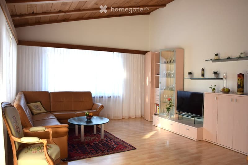 Living room with a brown leather sofa, a wooden armchair, a round coffee table, a television on a cabinet, shelves with decorative items, and a vase with flowers on the coffee table.
