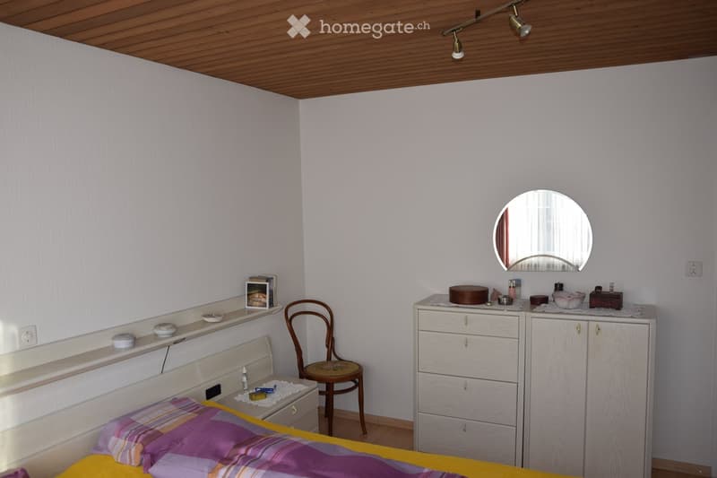Cozy bedroom with a wooden bed, colorful bedding, a wooden chair, a vanity cabinet with a mirror, and various personal items on shelves and surfaces.