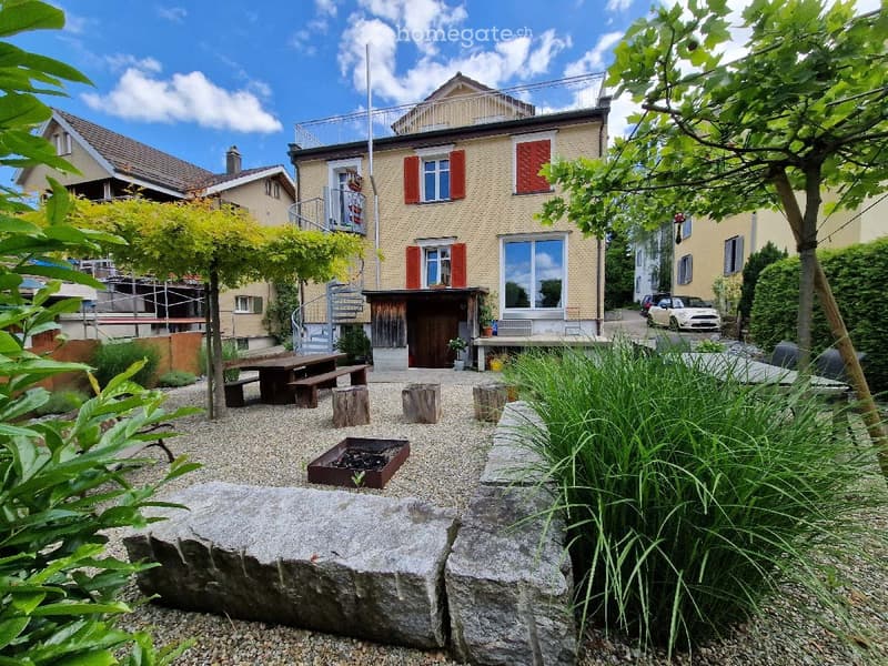 A well-maintained garden area with a wooden bench, stone seating, a fire pit, and lush greenery, adjacent to a two-story house with red shutters and a balcony.