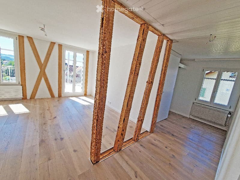 Empty room with wooden flooring, exposed wooden beams, large windows with a view, and white walls.
