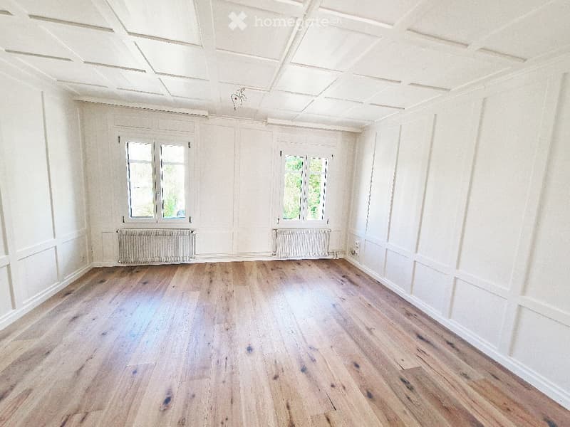 Empty room with wooden flooring, white walls, and large windows with radiators beneath them.