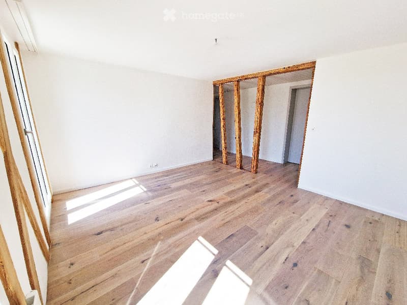 Empty room with light wooden flooring and white walls, featuring exposed wooden beams and a doorway leading to another space.