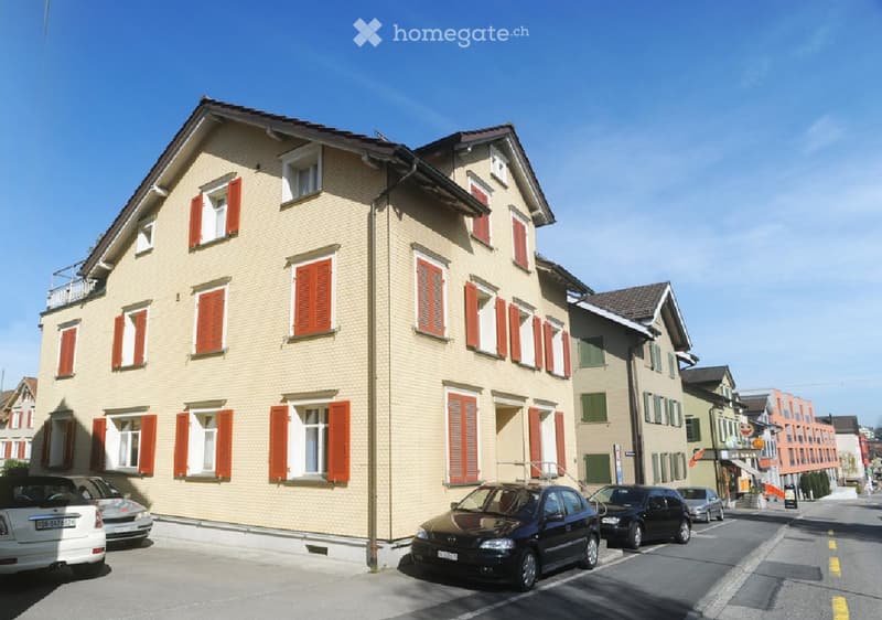 A two-story residential building with beige brick walls, red shutters, and multiple windows. The building has a sloped roof and is situated on a street with parked cars.