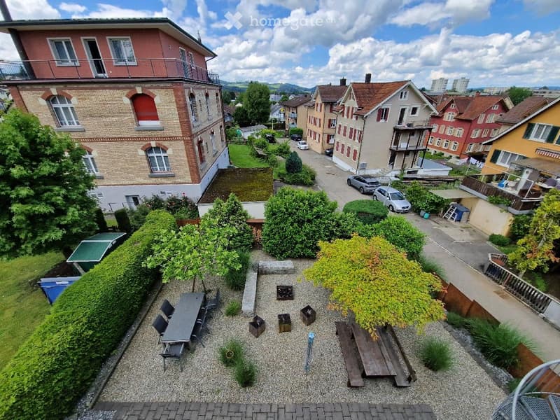A well-maintained garden area with a gravel pathway, a wooden bench, a small table with chairs, and several potted plants and shrubs. The garden is surrounded by hedges and is part of a residential neighborhood with multi-story houses and parked cars visi