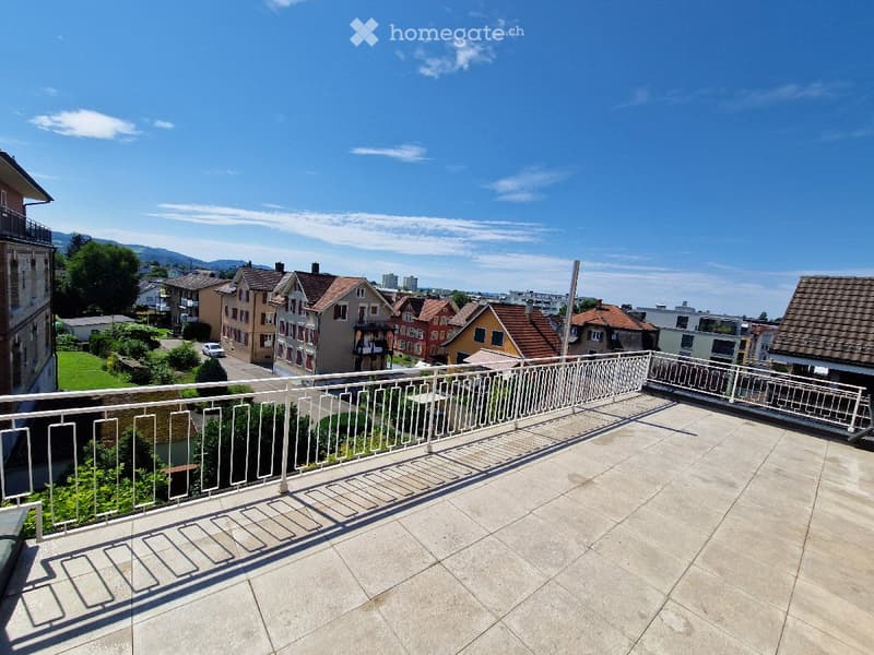 Balcony with white metal railing, tiled floor, overlooking a residential area with houses, trees, and distant mountains.