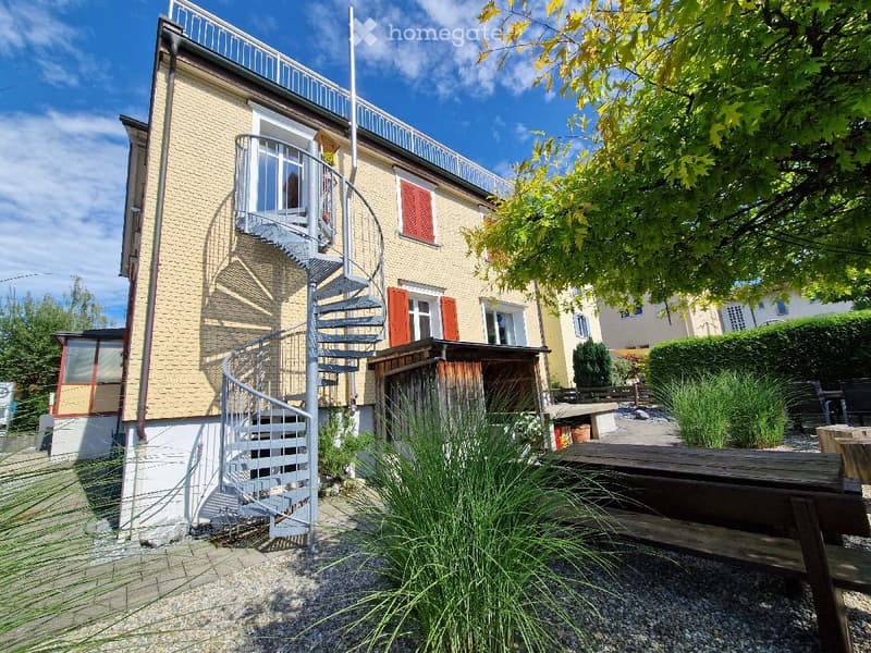 Two-story house with yellow brick facade, red shutters, spiral staircase leading to a balcony, and a small outdoor seating area with a wooden table and chairs.