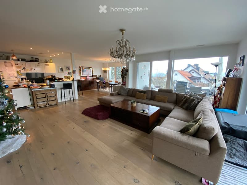 Open-plan living and dining area with wooden flooring, a large sectional sofa, coffee table, dining area with chairs, and a modern kitchen with white cabinets and stainless steel appliances.