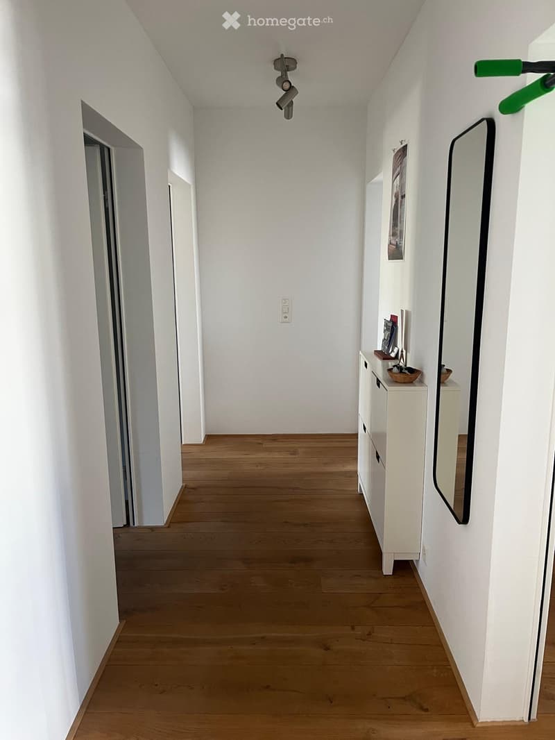 Clean, minimalist hallway with light wooden flooring, white walls, and a small white cabinet with a mirror and decorative items on top.