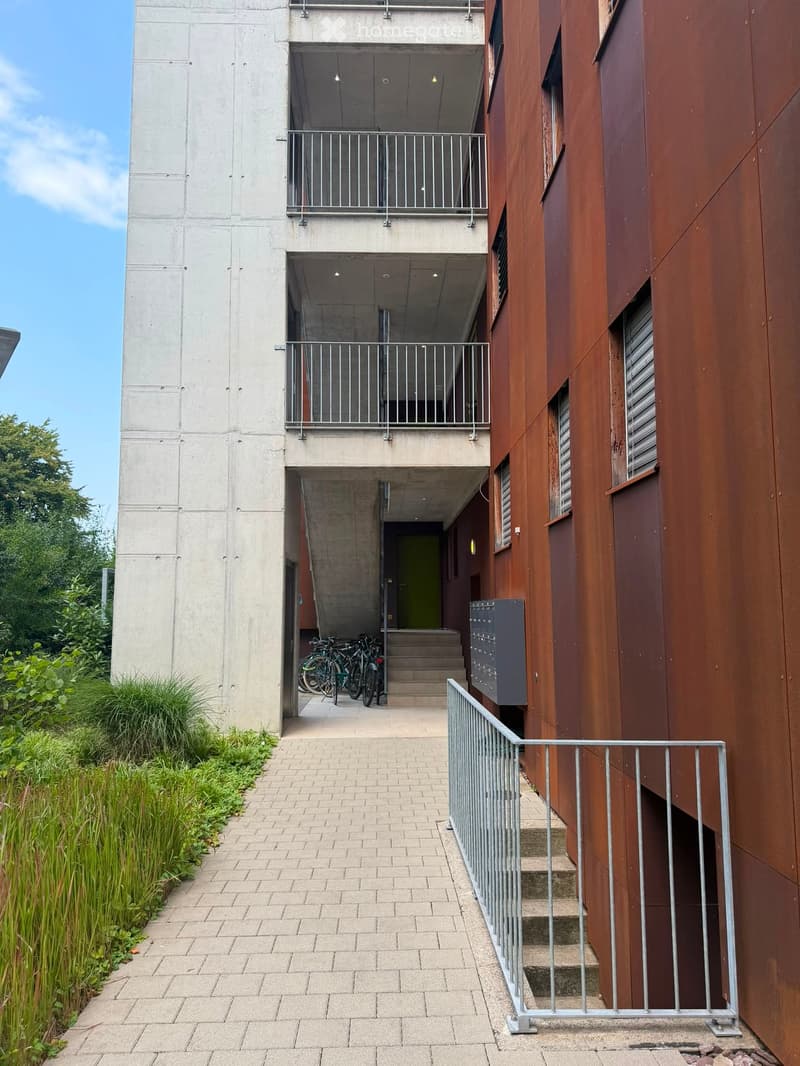 Modern multi-story building with concrete and wooden facades, featuring balconies, bicycles parked near the entrance, and a landscaped pathway.