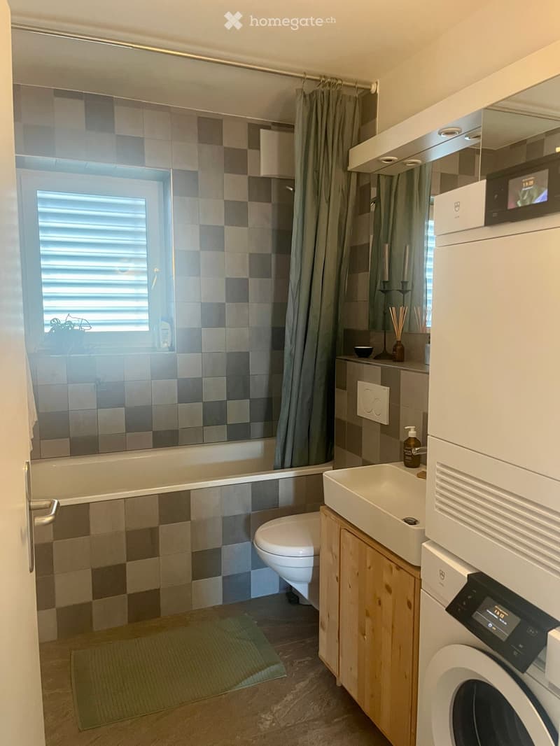 Modern bathroom with a bathtub, toilet, sink, and washing machine. Features a tiled shower area with a curtain, a wooden cabinet, and a small window with blinds.