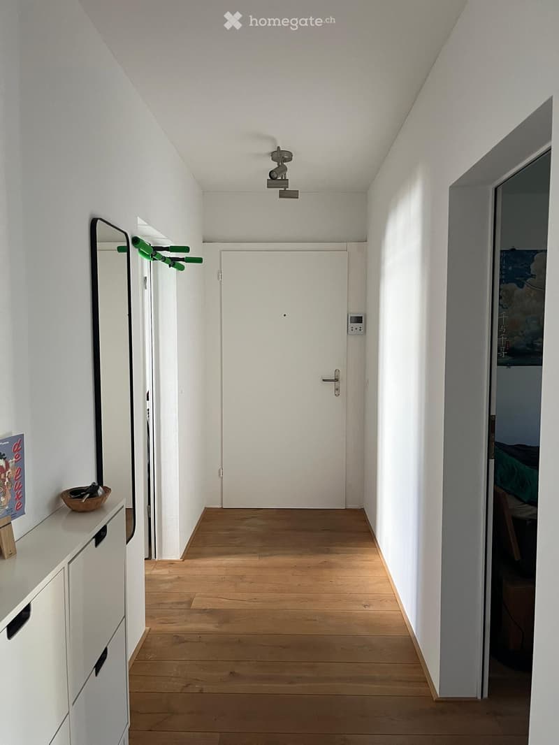 A narrow hallway with light wooden flooring, white walls, and a white door at the end. A small white cabinet with a drawer is on the left, and a mirror is mounted on the wall above it. A small basket with keys sits on the cabinet.