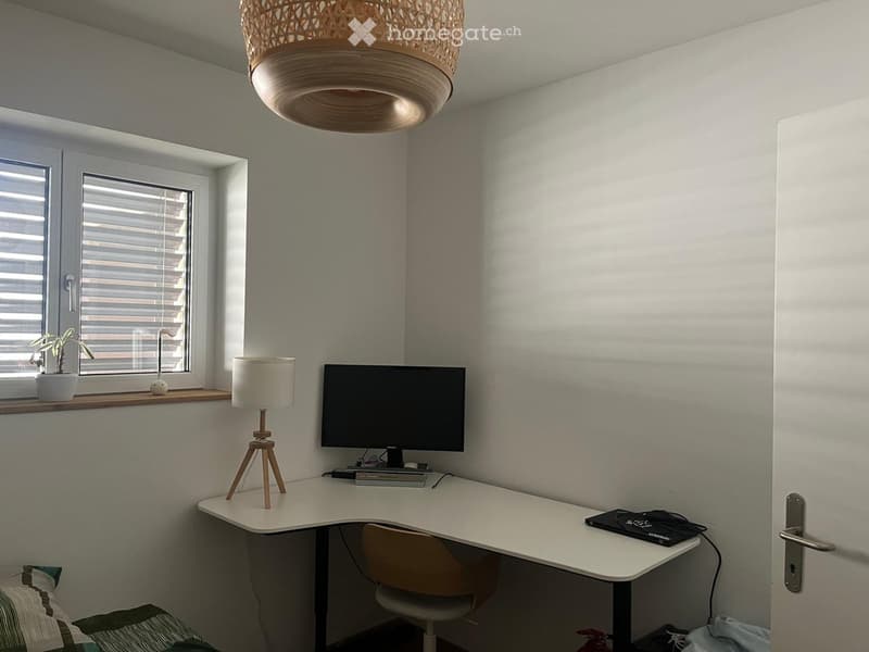 A small, modern room with a desk, chair, lamp, screen, and a plant on the windowsill. The room features a hanging woven light fixture and a bed with striped bedding visible in the corner.
