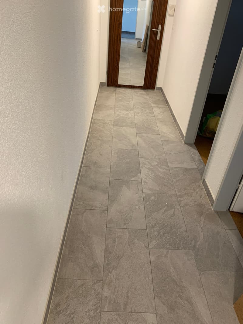 Clean hallway with tiled floor and white walls, featuring a wooden door with a mirror and a partially visible room on the right.
