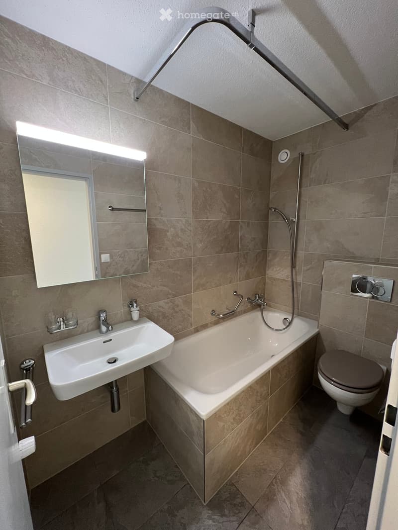 Modern bathroom with beige tiles, a white sink, bathtub, toilet, and a large mirror with built-in lighting. Features a handheld showerhead and a grab bar for safety.