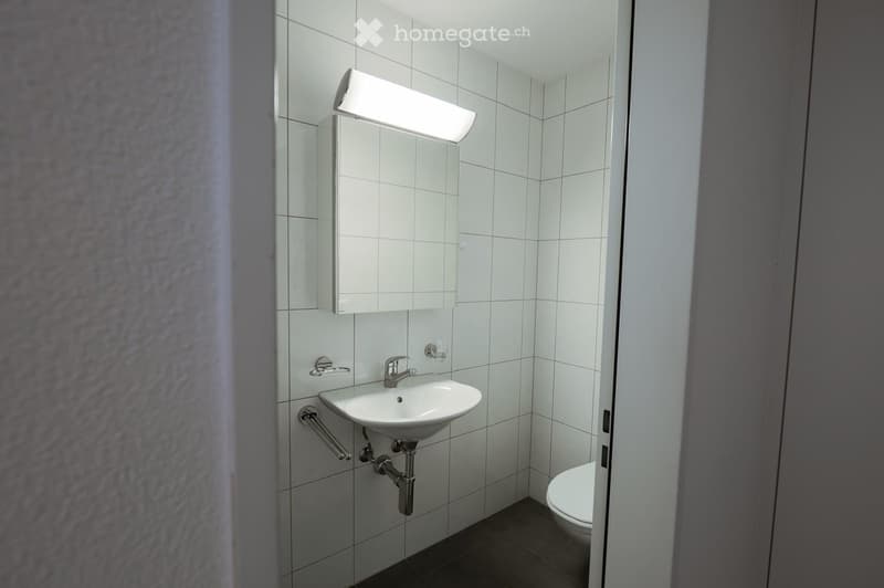Modern bathroom with white tiled walls, a white sink with a chrome faucet, a mirror with a built-in cabinet, and a toilet. The space is compact and well-lit with a fluorescent light fixture above the mirror.
