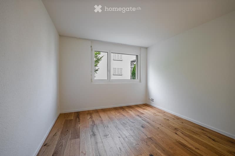Empty room with light wooden flooring and white walls, featuring a large window with a view of greenery and a neighboring building.