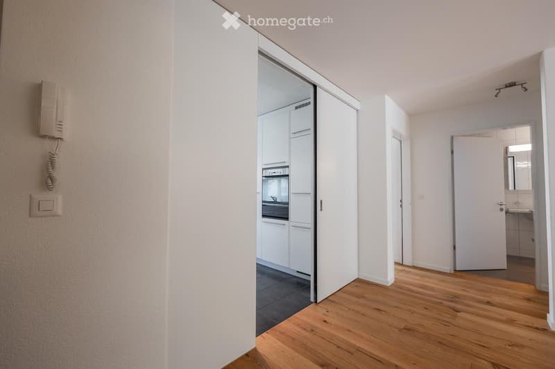 Empty room with wooden flooring, white walls, and open doors leading to a kitchen and bathroom. No furniture or equipment is present.