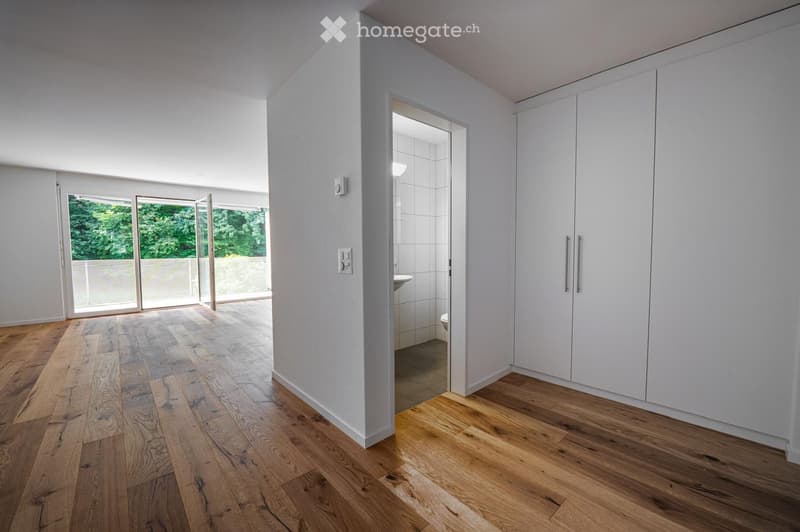 Empty room with wooden flooring, white walls, large sliding glass doors leading to an outdoor area, and a view of greenery.
