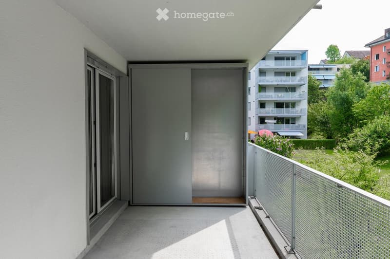 Modern balcony with a sliding glass door, metal railing, and a view of a green garden and neighboring buildings.