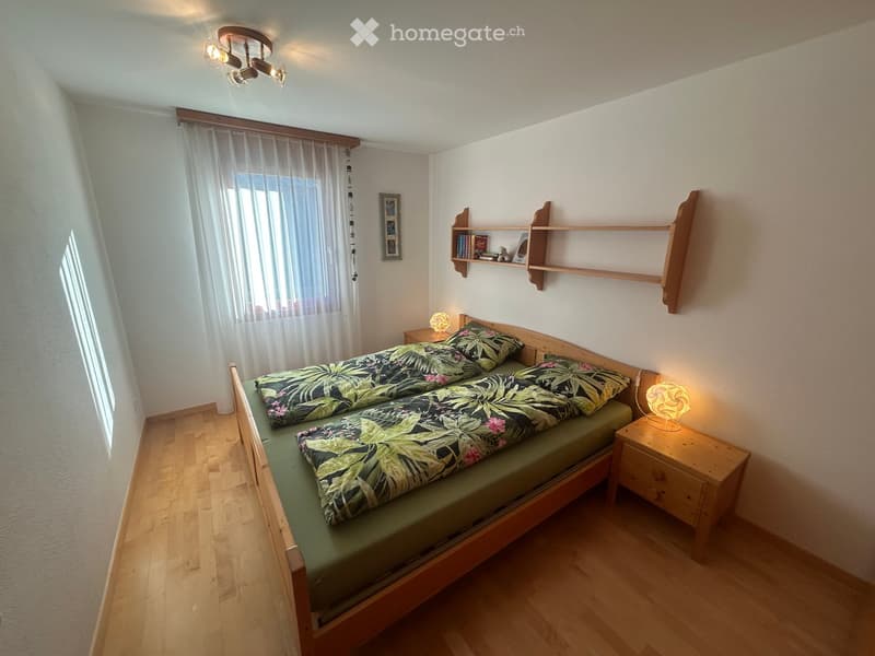 Bedroom with two beds, white walls, wooden floor, window with white curtains, wooden nightstands, and shelves with books and decor