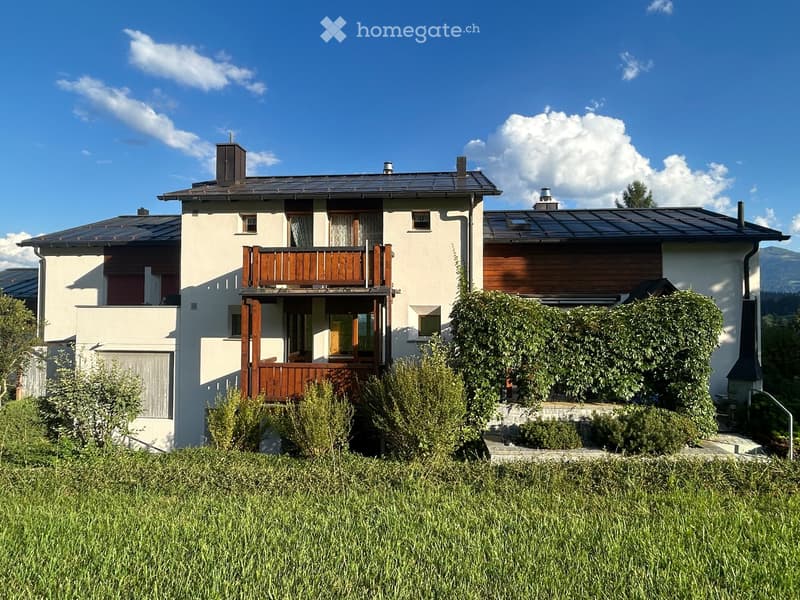 Two-story house with white walls, wooden balcony, and solar panels on the roof, surrounded by greenery and a grassy lawn.