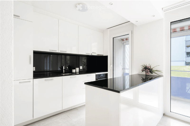 Modern white kitchen with sleek cabinets, black countertops, and a central island. Features a sink, faucet, and a small flower vase on the island.