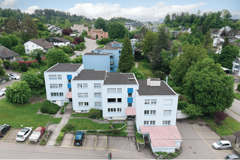 Aerial view of a multi-story residential building with light blue walls and white trim, surrounded by greenery and parked cars in a parking lot.