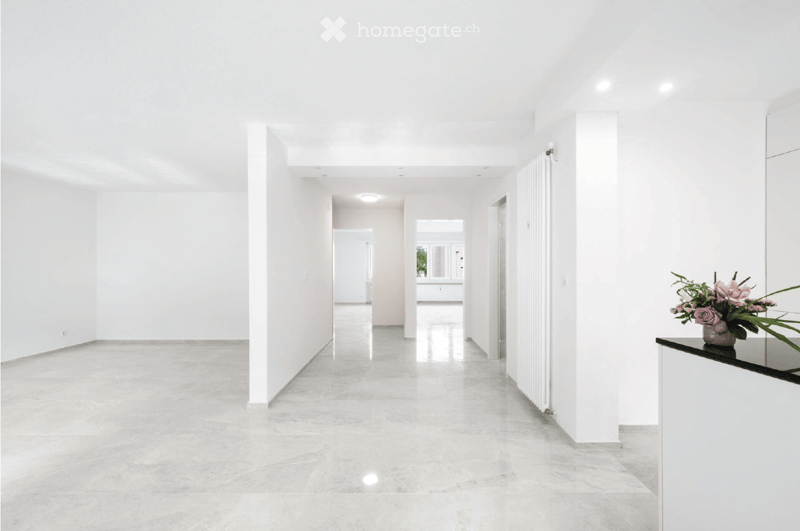 Bright, minimalist hallway with white walls, polished marble floors, and recessed lighting. Features a small vase with pink flowers on a black console table to the right.