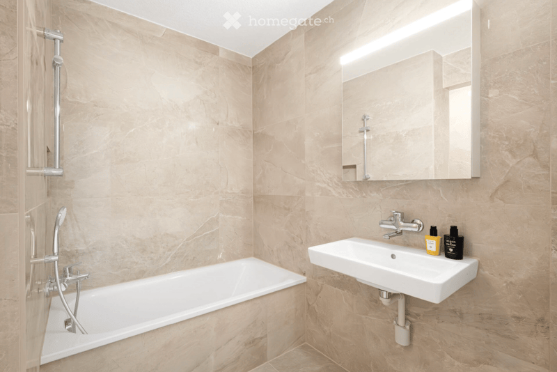 Modern bathroom with marble walls and floor, featuring a bathtub, a sink with a mirror, and a shower head.