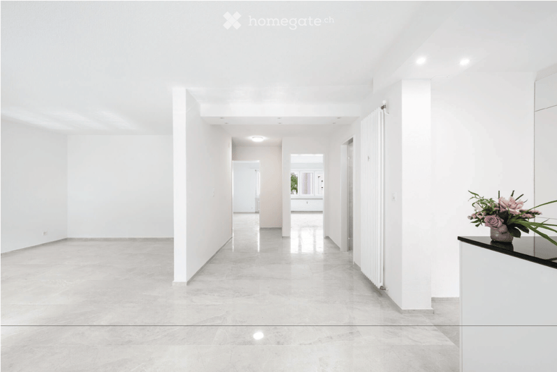 Bright, minimalist hallway with white walls, polished marble floors, and recessed lighting. Features a small black console table with a vase of pink flowers on the right side.