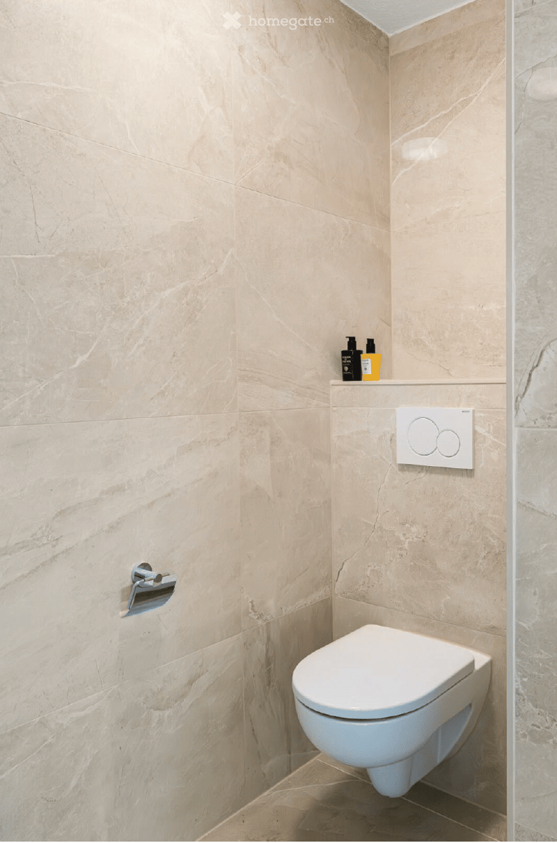 Modern bathroom with beige marble tiles, a toilet, a wall-mounted faucet, a control panel, and toiletries on a shelf.