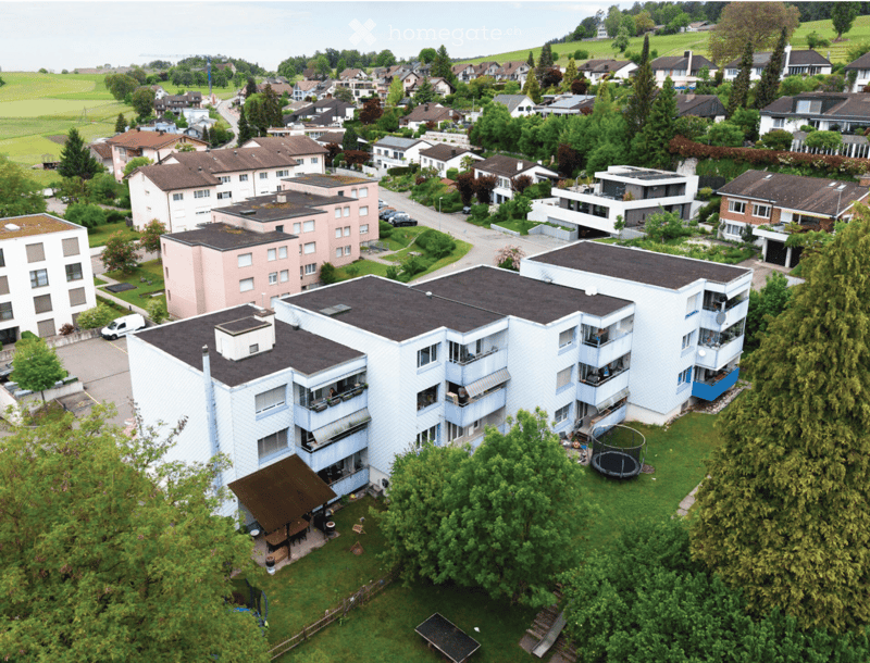 Aerial view of a multi-story residential building surrounded by greenery, with balconies, a playground, and nearby houses.