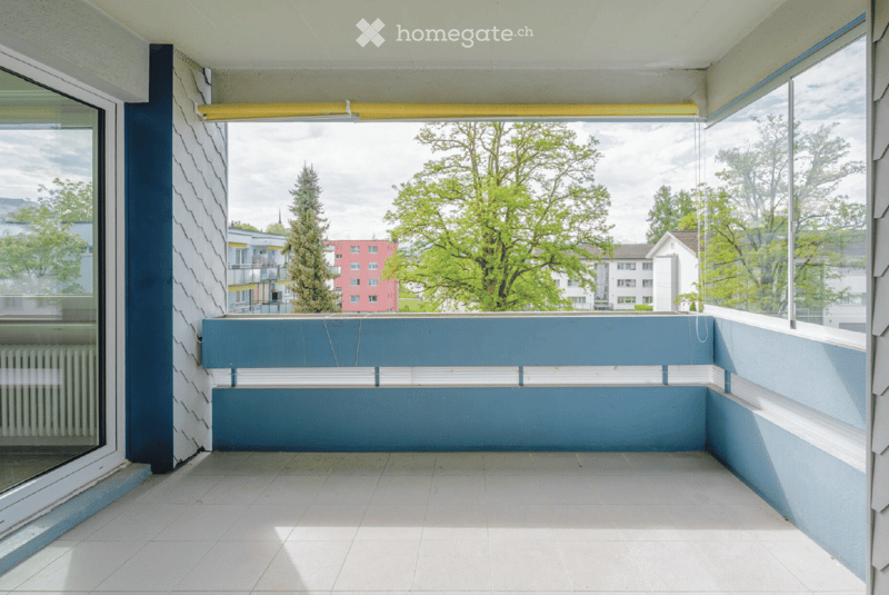 Modern balcony with blue walls, white tiled floor, large windows offering a view of trees and buildings.