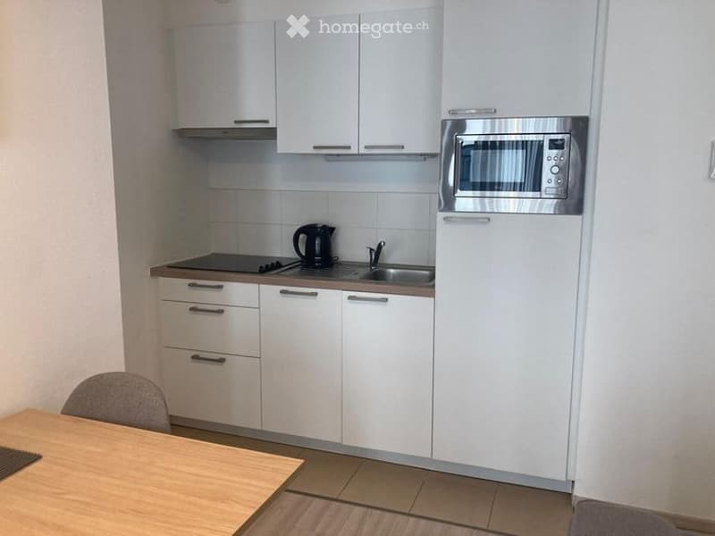 Modern kitchen with white cabinets, stainless steel microwave, refrigerator, stove, sink, and a small dining area with a table and chair.
