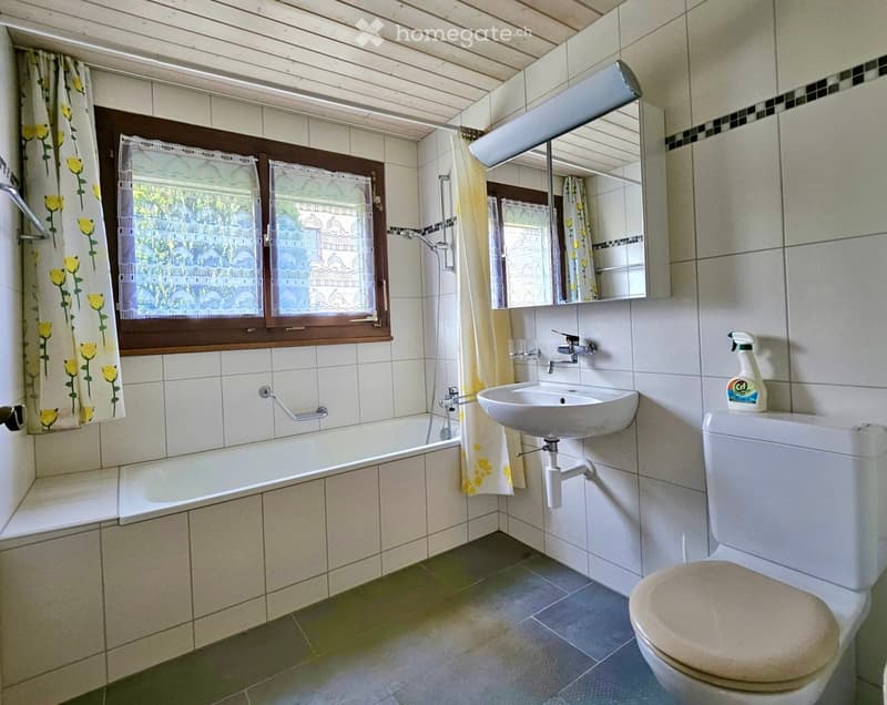Bathroom with a bathtub, sink, toilet, mirror, and window with floral curtains.