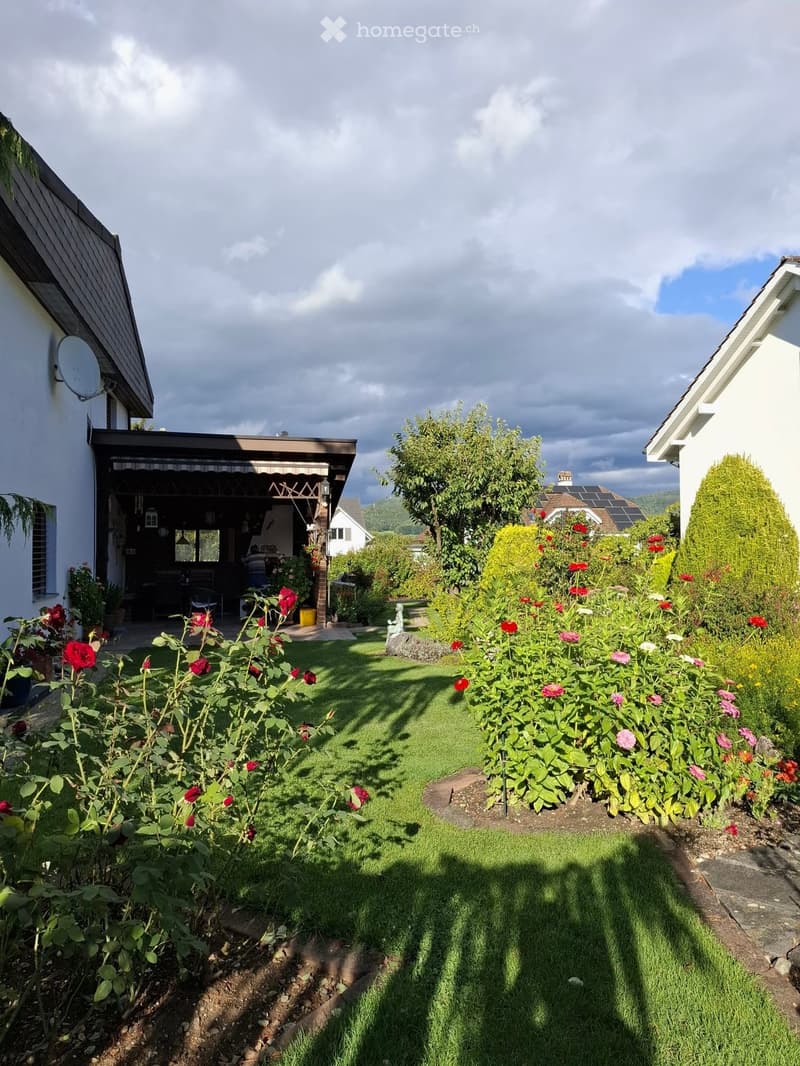 Well-maintained garden with a variety of colorful flowers, neatly trimmed grass, and a covered outdoor seating area connected to a house.