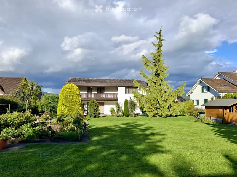 Well-maintained garden with a large, neatly trimmed lawn, surrounded by trees and shrubs, with a white house featuring a balcony in the background.