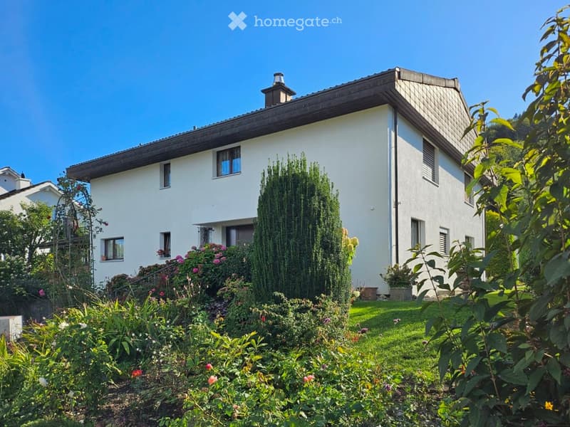 A two-story white house with a modern design, surrounded by a well-maintained garden with various plants and flowers.