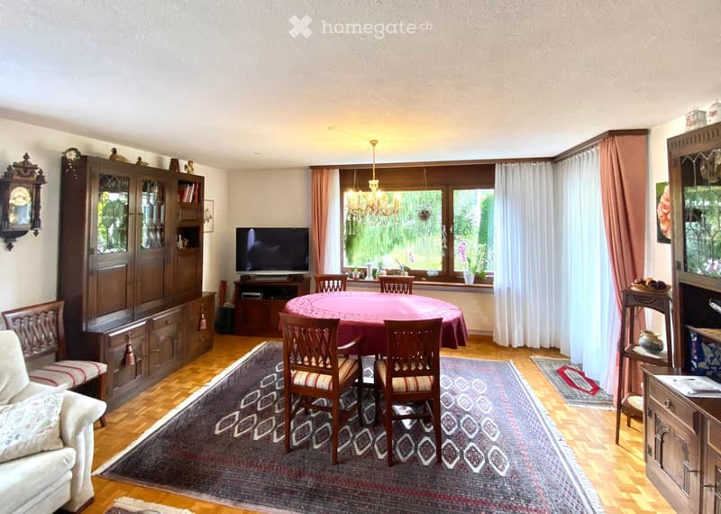 A well-furnished room with wooden flooring, a large rug, a sofa, a dining table with chairs, a television, and wooden cabinets.